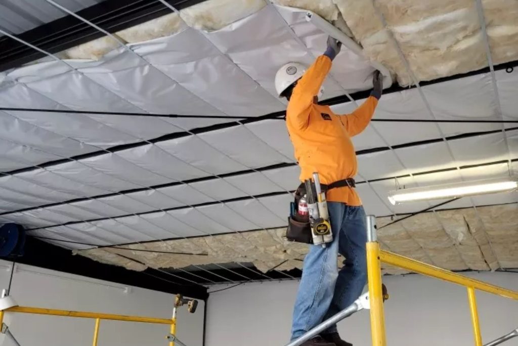 A man works on the ceiling of a warehouse, standing on a ladder and focused on his task.