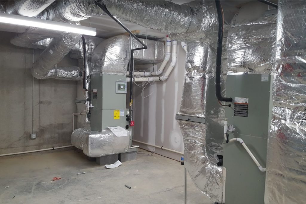 An industrial HVAC room with two large units and extensive insulated ductwork on concrete walls and floor.