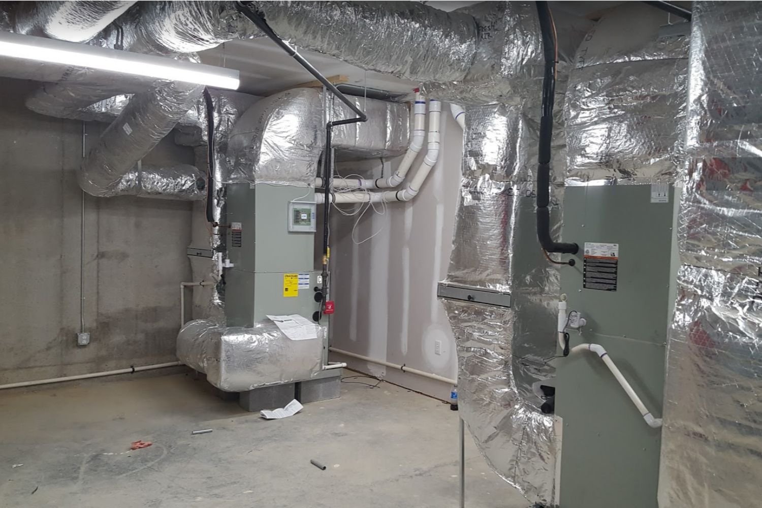 An industrial HVAC room with two large units and extensive insulated ductwork on concrete walls and floor.