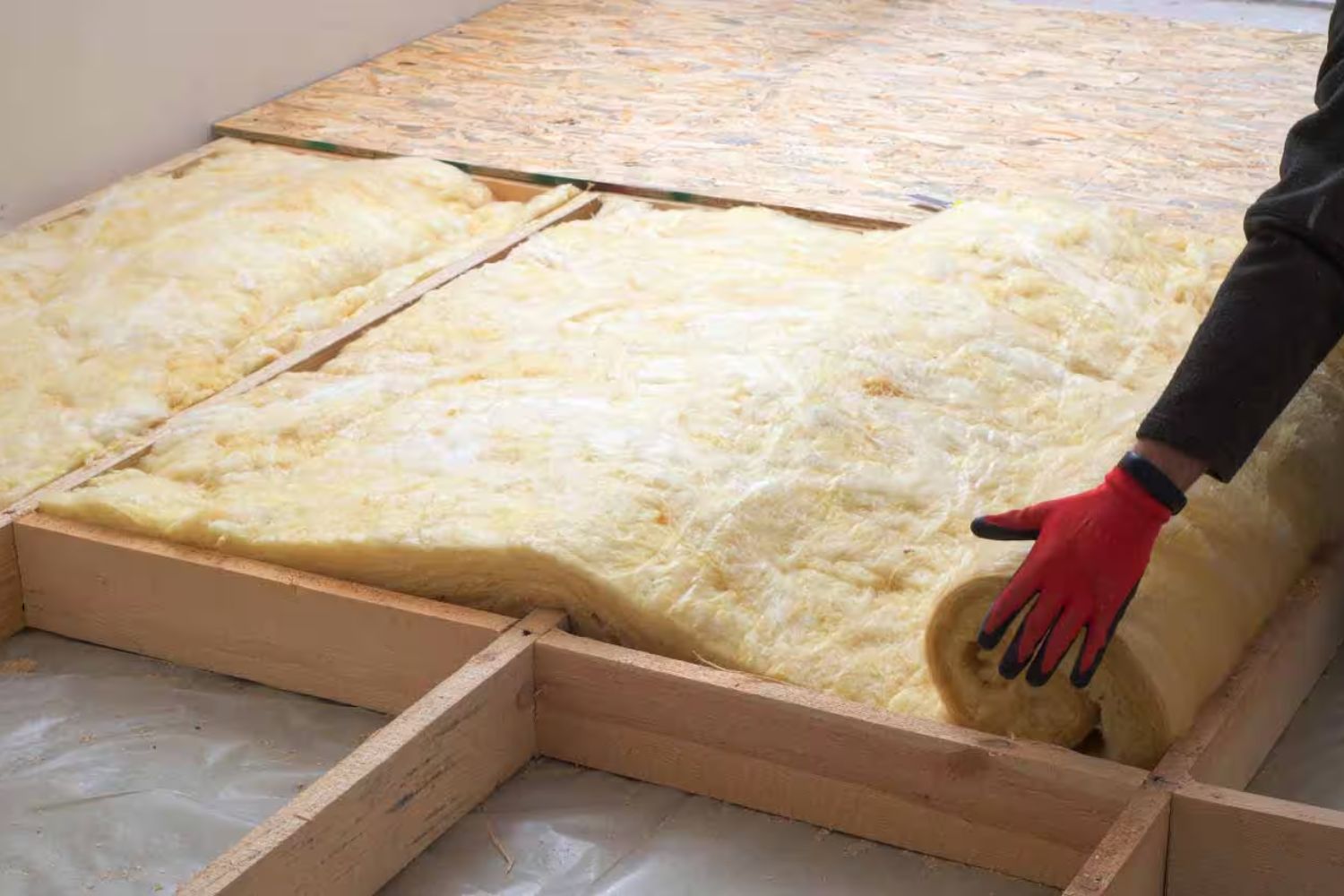 A person wearing a red glove installs yellow insulation between wooden beams on a floor, preparing for construction or renovation.