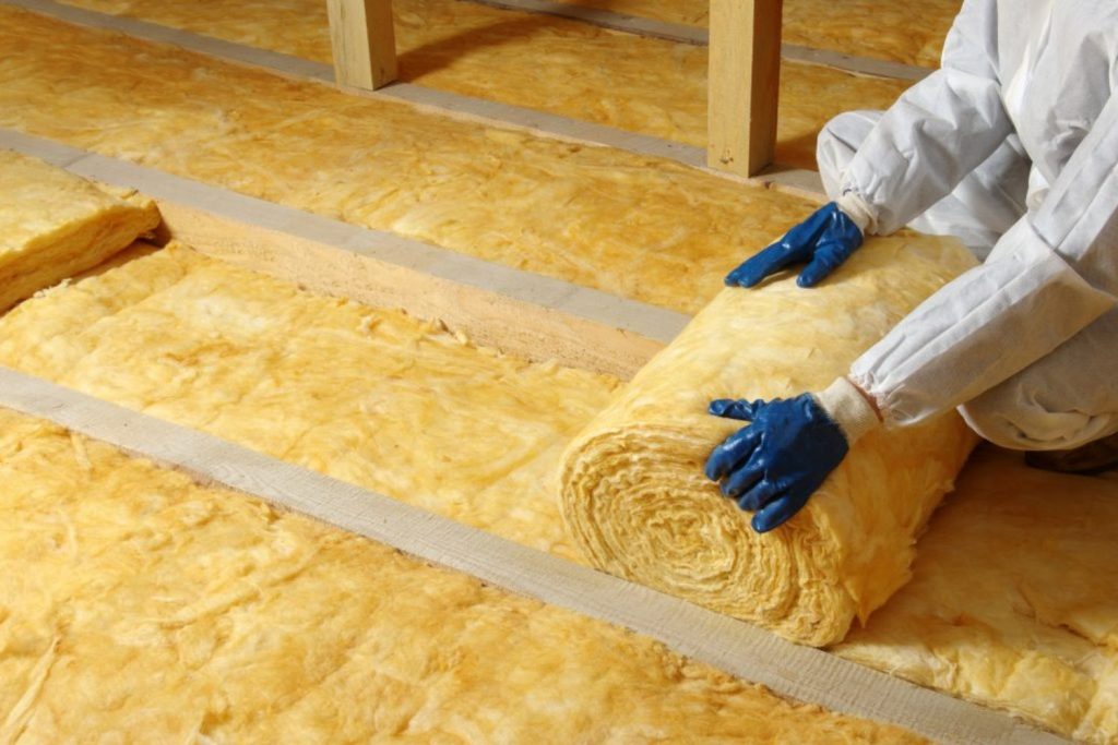 A man in a white suit and blue gloves installs insulation on the floor, focusing on his task with precision.