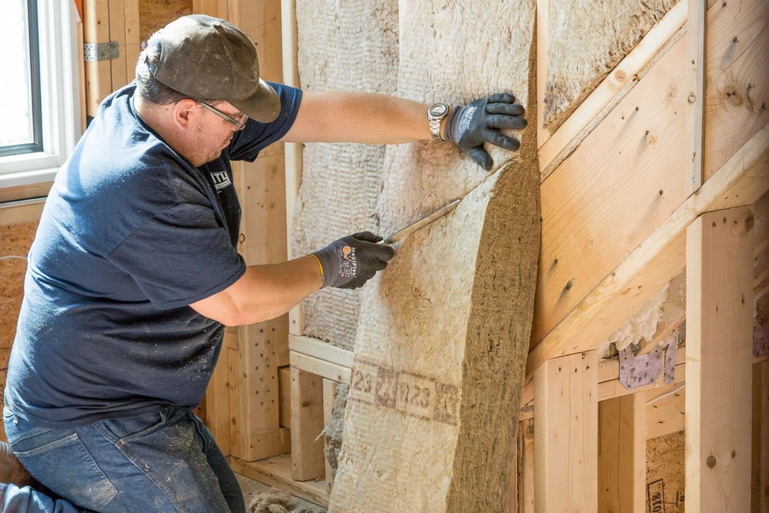 A man installs insulation in a wooden frame, cutting the material with a utility knife at a well-lit construction site.